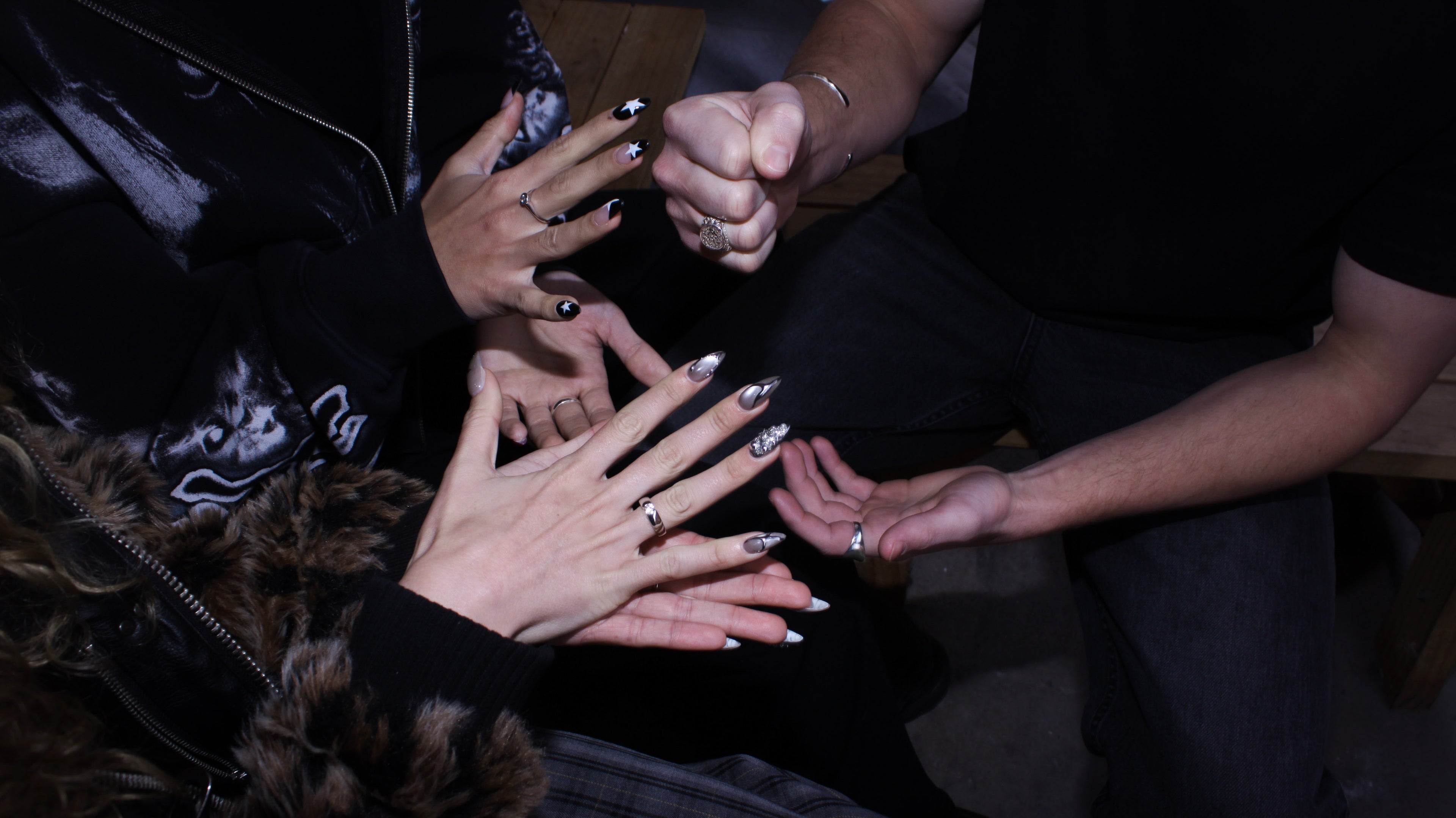 Close-up of multiple hands gathered together, each wearing long black manicured nails with silver tips and small silver ring accents, photographed in dim indoor lighting.